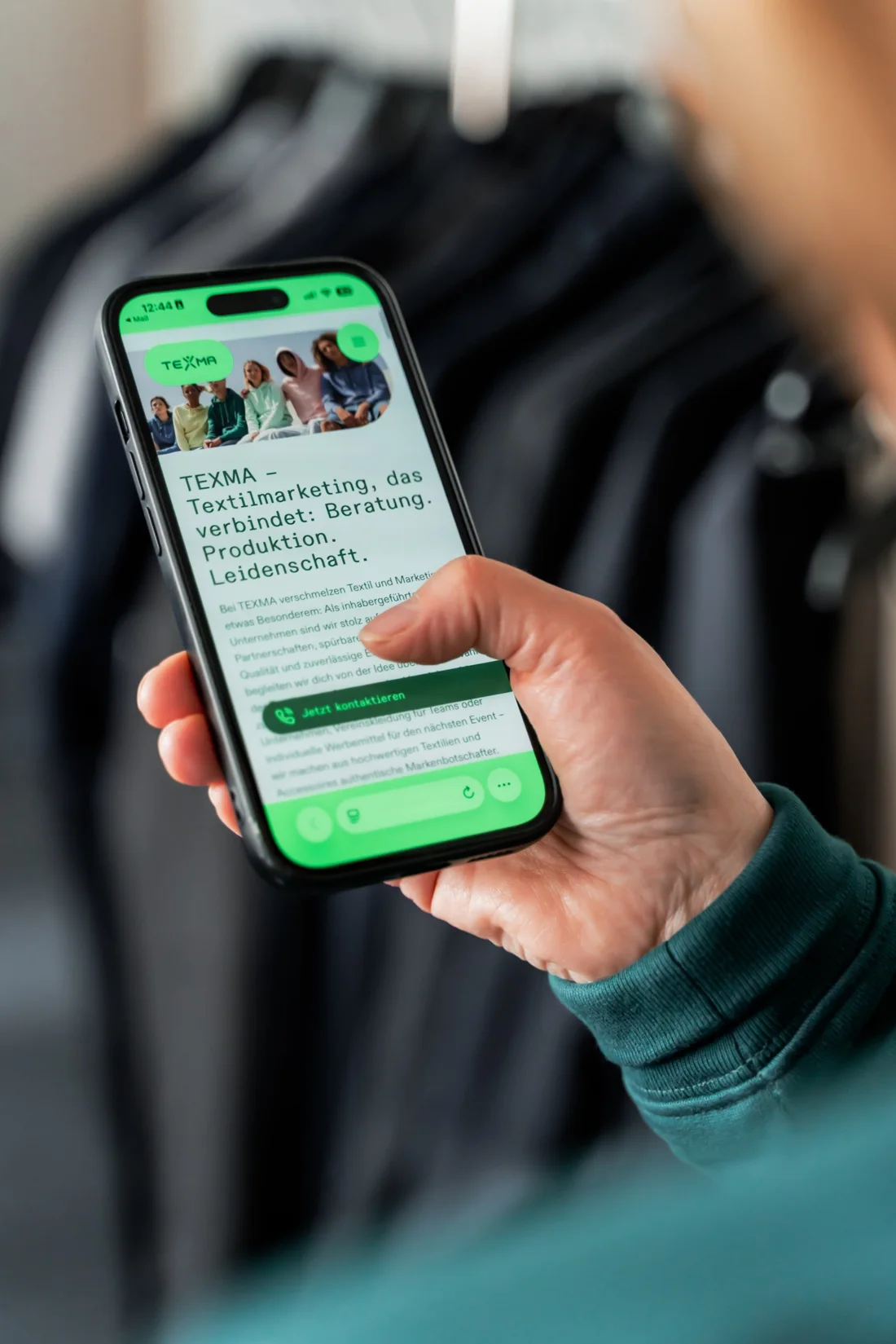 Hand hält ein Smartphone mit der TEXMA-Website vor einer Kleiderstange